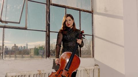 Eine Frau steht mit ihrem Cello in Abendkleidung in einem Industriegebäude mit großen Fenstern und altmodischem Heizungskörper.