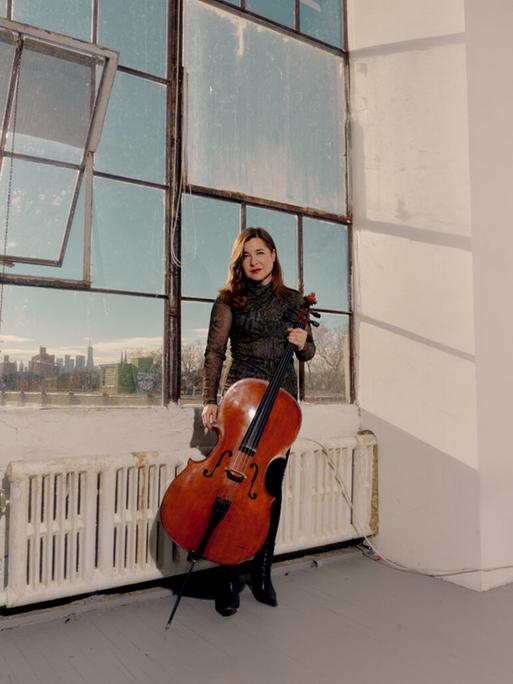 Eine Frau steht mit ihrem Cello in Abendkleidung in einem Industriegebäude mit großen Fenstern und altmodischem Heizungskörper.