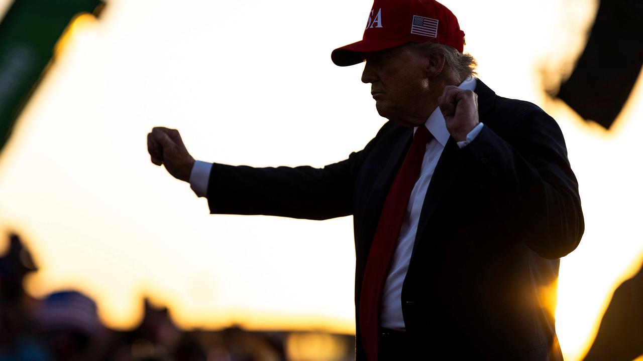 US-Präsident Donald Trump trägt ein rotes Cap mit US-Flagge an der Seite und steht auf einer Bühne mit erhobenen Armen und geballten Fäusten 