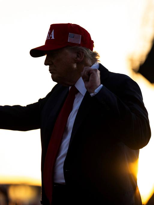US-Präsident Donald Trump trägt ein rotes Cap mit US-Flagge an der Seite und steht auf einer Bühne mit erhobenen Armen und geballten Fäusten 