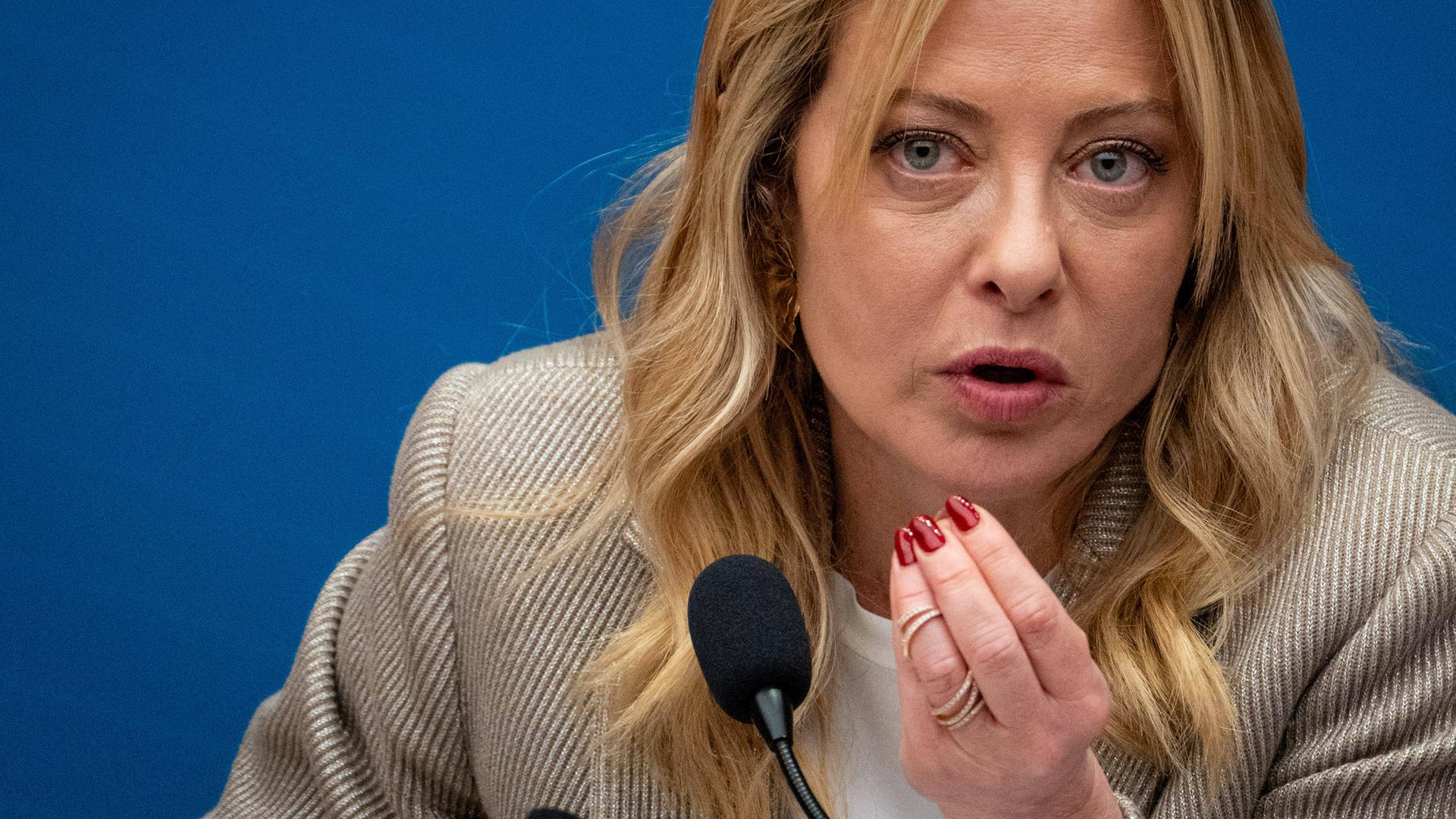 Giorgia Meloni, die Premierministerin Italiens, sitzt bei einer Pressekonferenz vor einem Mikrofon. Sie spricht und hält dabei die Hand in die Luft.