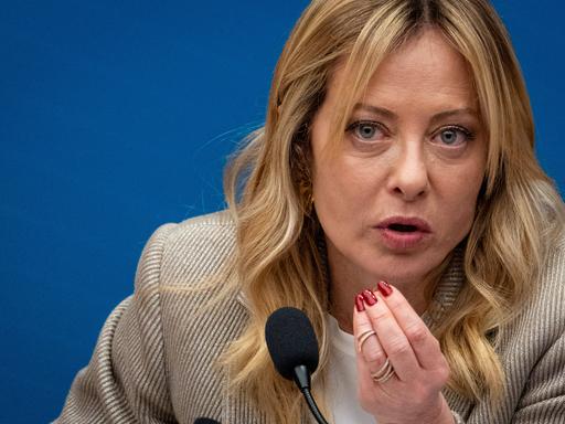 Giorgia Meloni, die Premierministerin Italiens, sitzt bei einer Pressekonferenz vor einem Mikrofon. Sie spricht und hält dabei die Hand in die Luft.