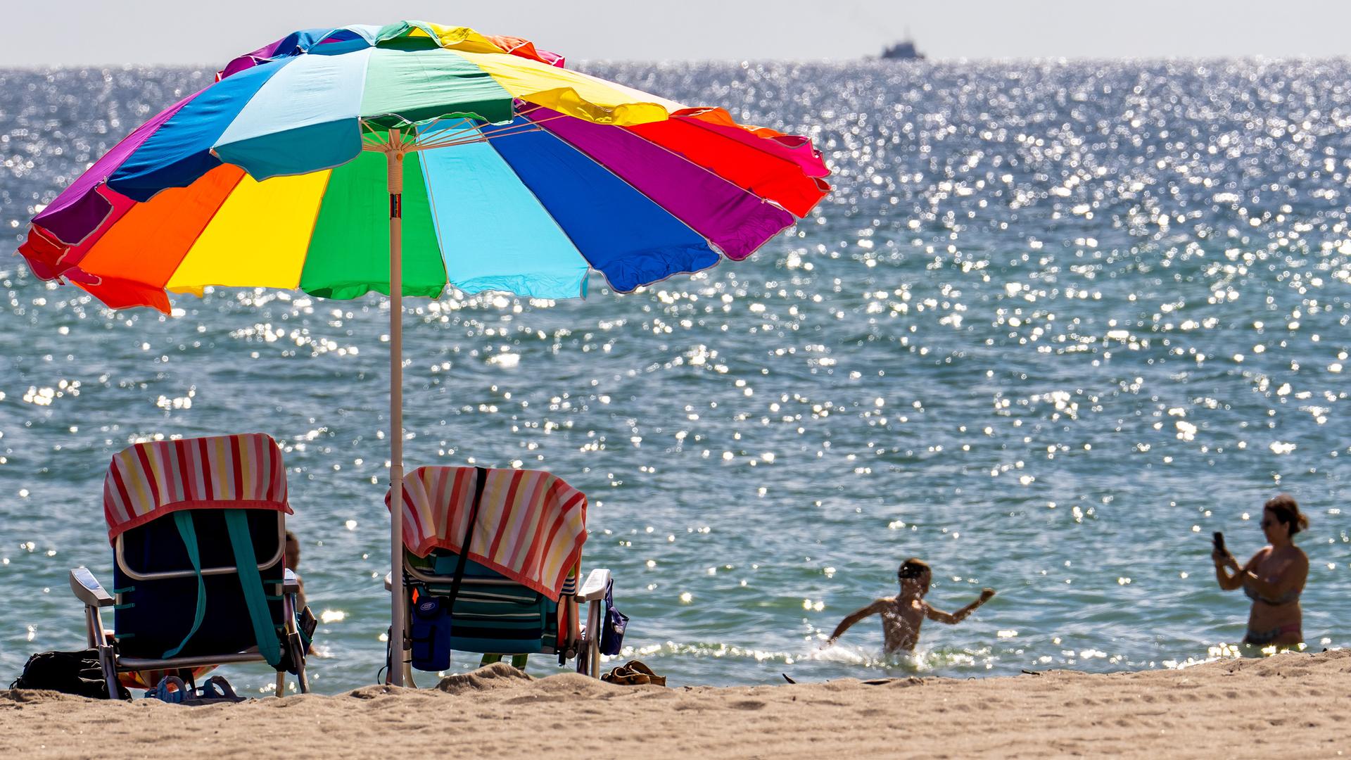 Ein bunter Sonnenschirm am Strand, im Meer planschen ein Kind und eine Frau