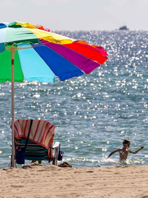 Ein bunter Sonnenschirm am Strand, im Meer planschen ein Kind und eine Frau