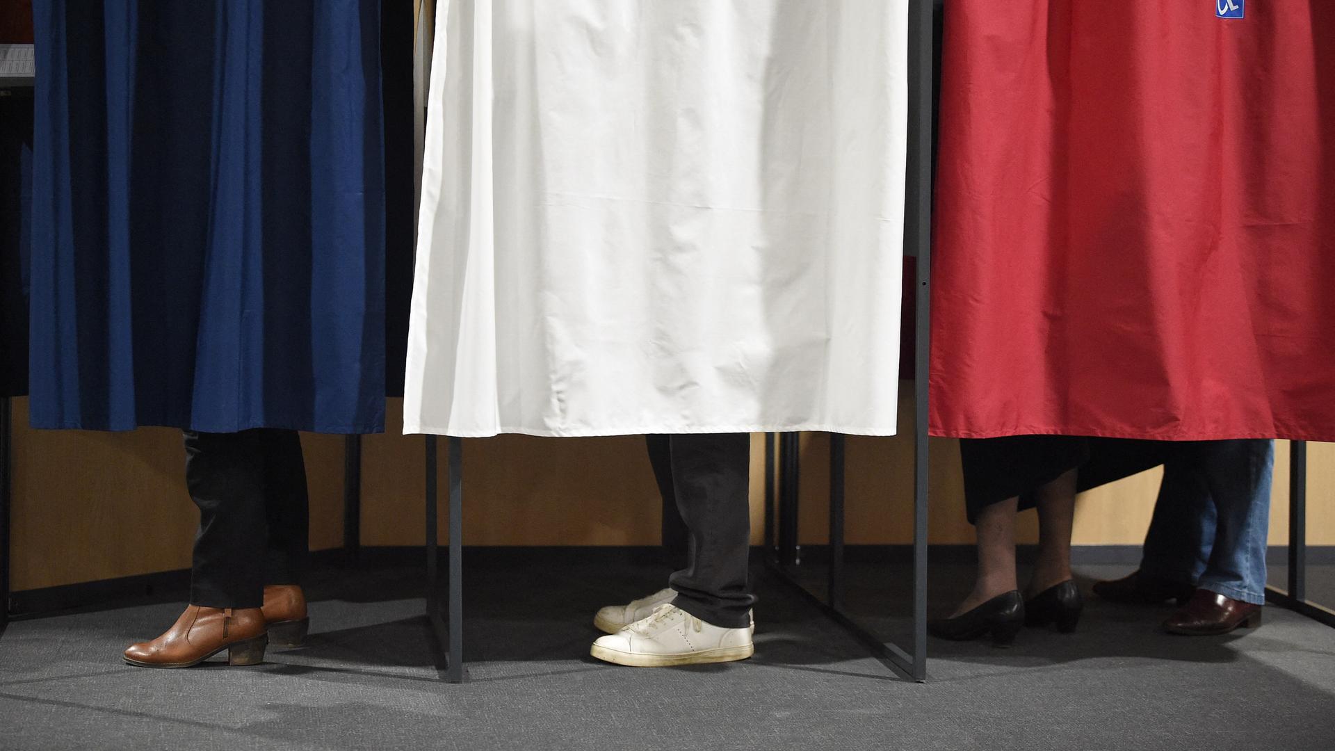 Eine Wahlkabine deren Vorhänge die Farben der Französischen Flagge zeigen. Hinter den Vorhängen sind die Füsse von Wählenden zu sehen.