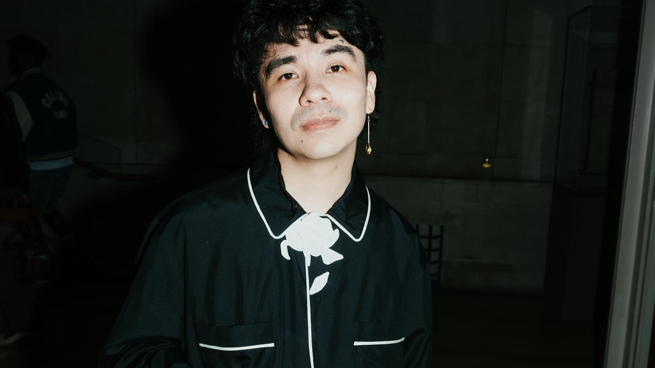 Ocean Vuong steht vor einem schwarzen Hintergrund, trägt ein schwarzes Hemd mit weißen Abnähern und schaut direkt in die Kamera