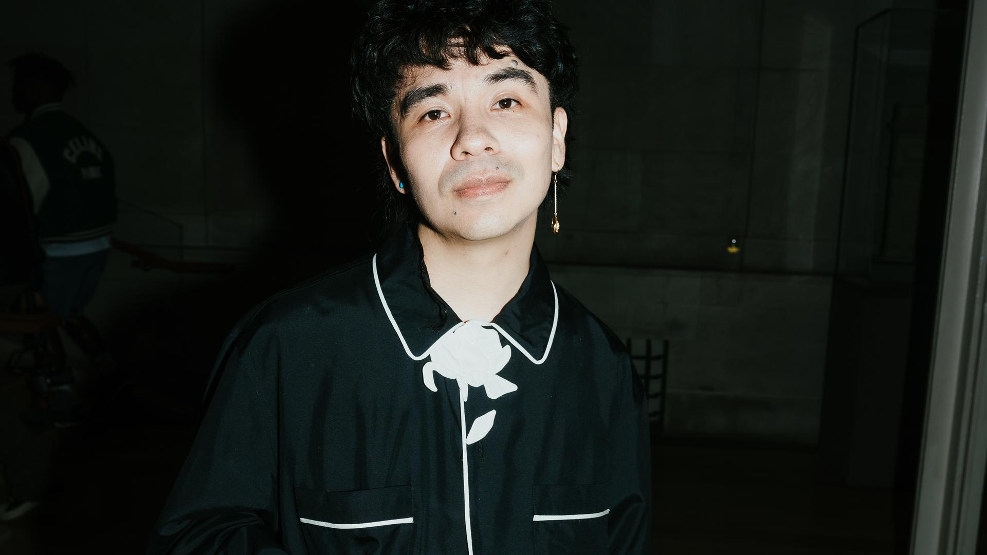 Ocean Vuong steht vor einem schwarzen Hintergrund, trägt ein schwarzes Hemd mit weißen Abnähern und schaut direkt in die Kamera