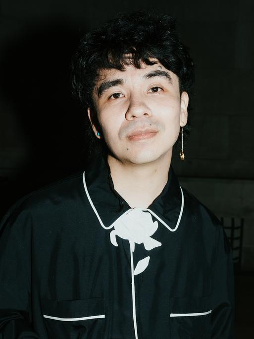 Ocean Vuong steht vor einem schwarzen Hintergrund, trägt ein schwarzes Hemd mit weißen Abnähern und schaut direkt in die Kamera