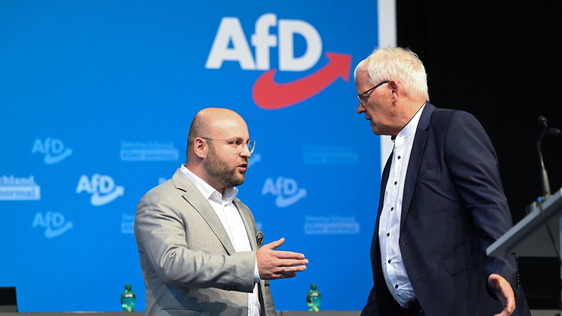 Die beiden stehen vor einer blauen Wand mit dem AfD-Logo und sprechen miteinander.
Die beiden stehen vor einer blauen Wand mit dem AfD-Logo und sprechen miteinander.