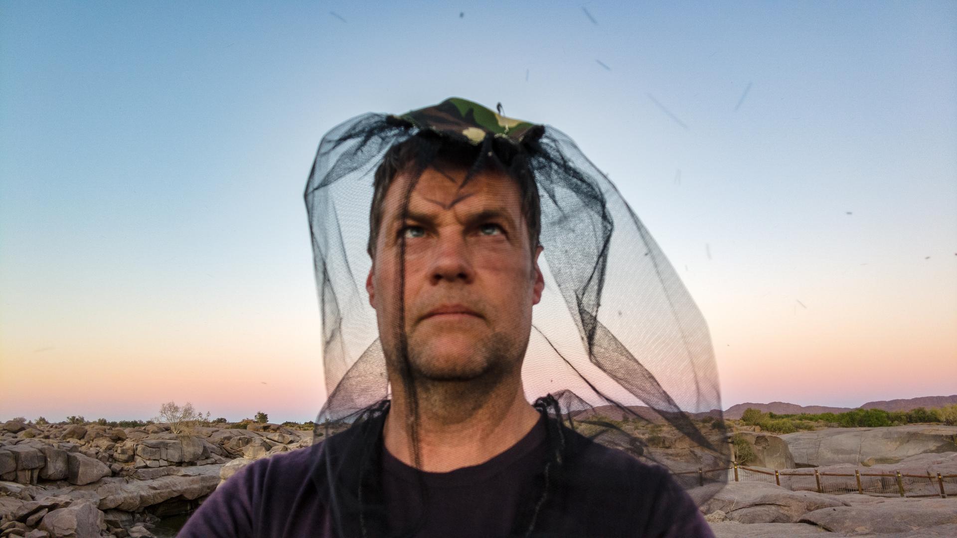 Ein Mann trägt ein Mückennetz über dem Kopf und schaut Richtung Himmel. Er steht am Meer.