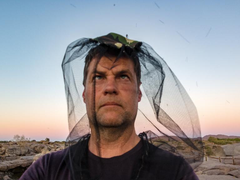 Ein Mann trägt ein Mückennetz über dem Kopf und schaut Richtung Himmel. Er steht am Meer.