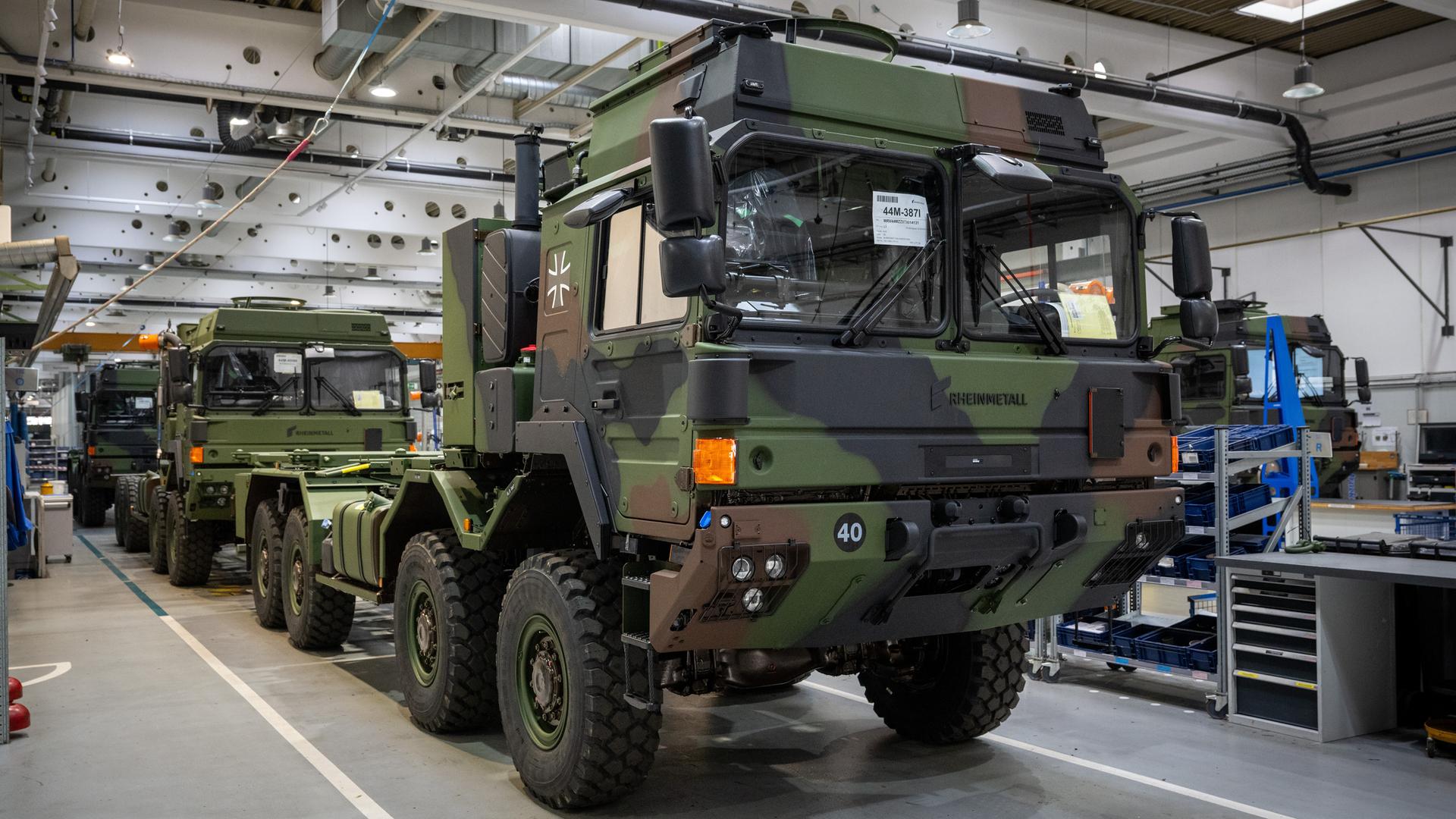 Militärische Lastwagen in Camouflage-Tarnfarben stehen in einer Fertigungshalle hintereinander. 
