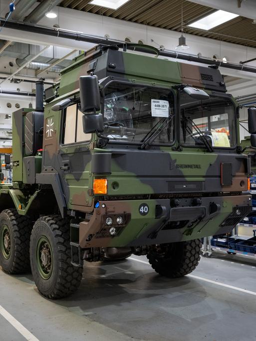 Militärische Lastwagen in Camouflage-Tarnfarben stehen in einer Fertigungshalle hintereinander. 