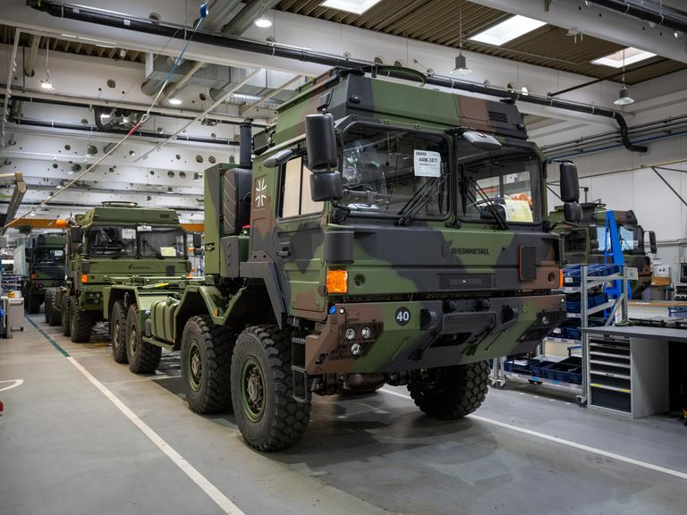 Militärische Lastwagen in Camouflage-Tarnfarben stehen in einer Fertigungshalle hintereinander. 