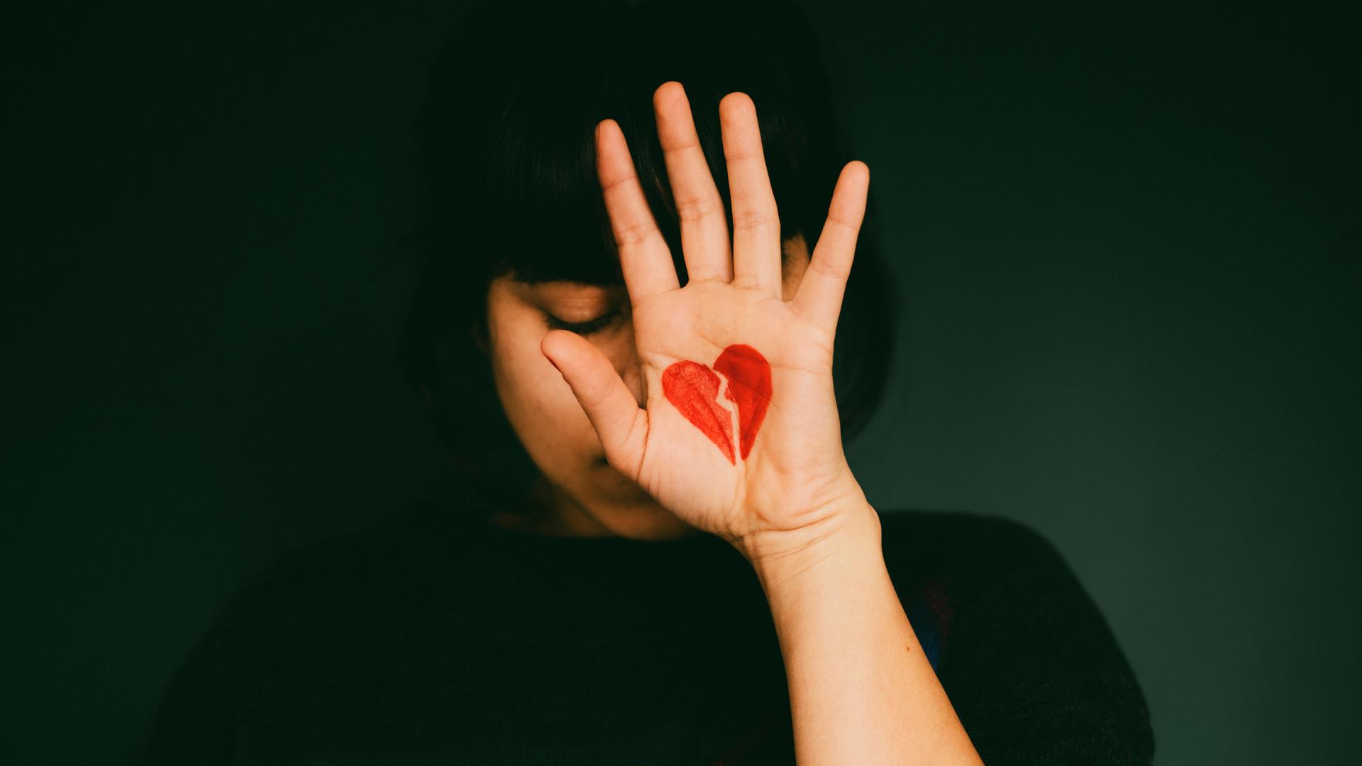 Eine Frau hält ihre Hand hoch. Auf ihrer Handfläche ist ein zerbrochenes Herz gemalt.