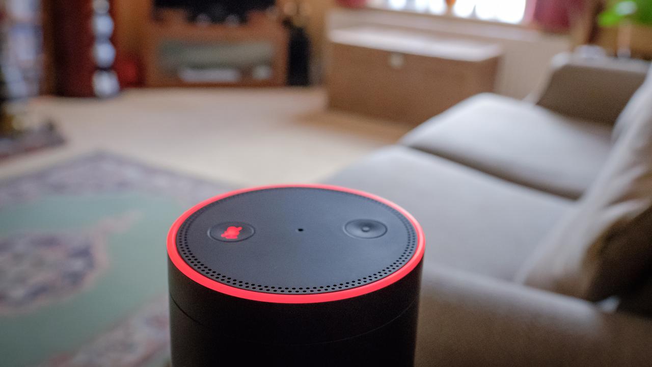 Der Lautsprecher eines Sprachassistenten steht in einem gepflegten Wohnzimmer, in dessen Hintergrund unter anderem ein Sofa, ein TV-Möbel und LED-Leuchten zu sehen sind.