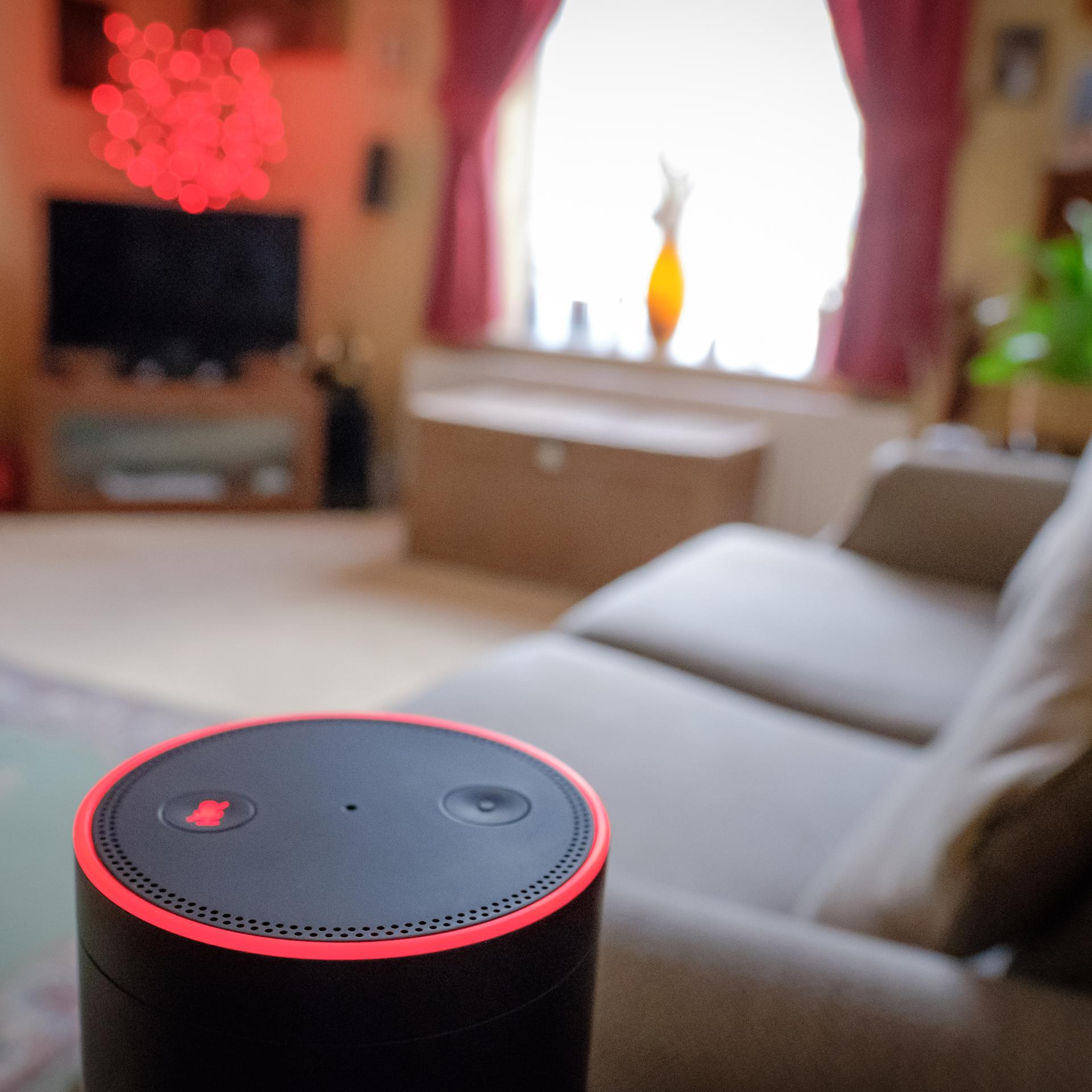 Der Lautsprecher eines Sprachassistenten steht in einem gepflegten Wohnzimmer, in dessen Hintergrund unter anderem ein Sofa, ein TV-Möbel und LED-Leuchten zu sehen sind.