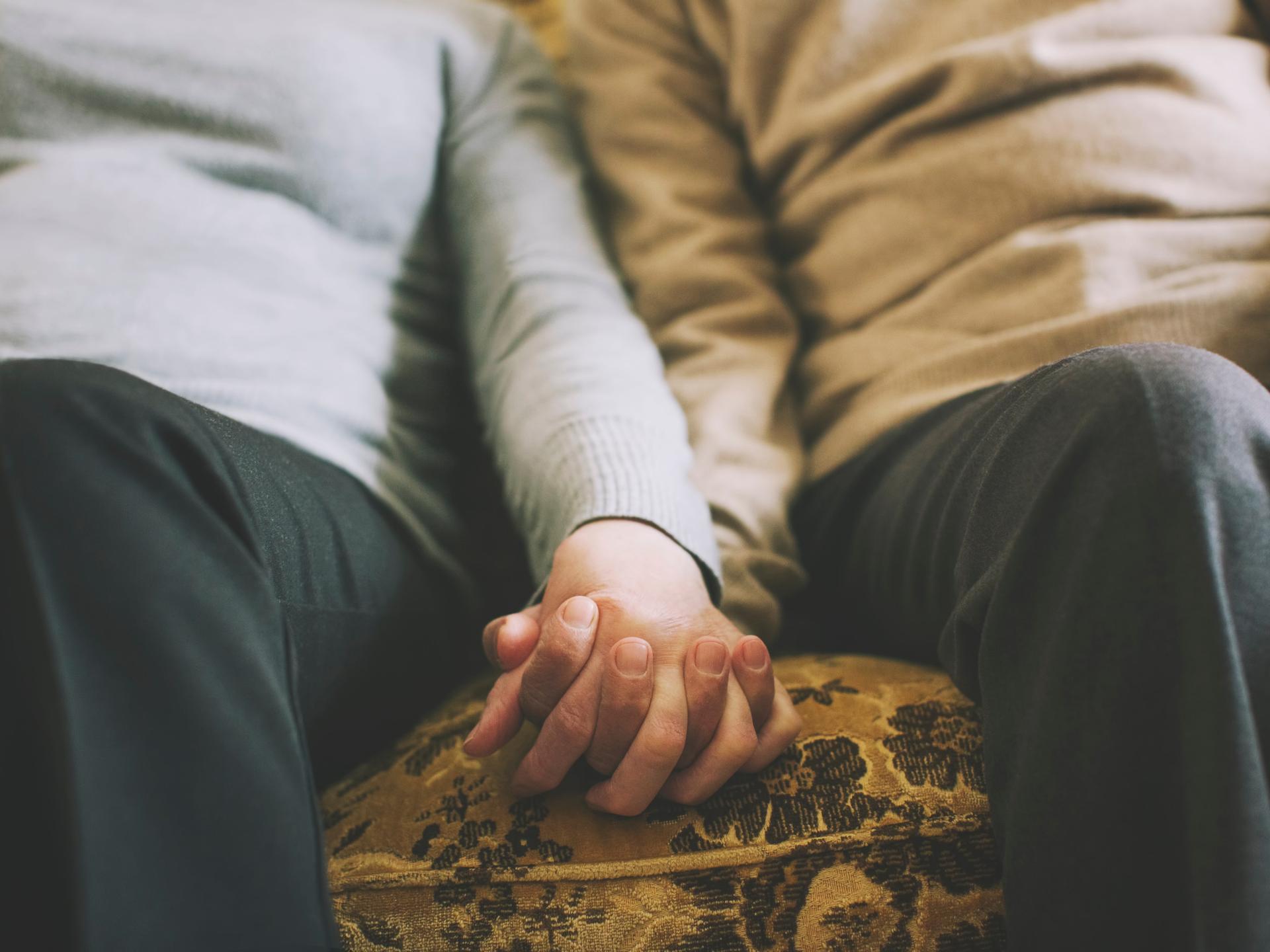 Zwei Menschen halten ihre Hände, während sie nebeneinander auf dem Sofa sitzen.