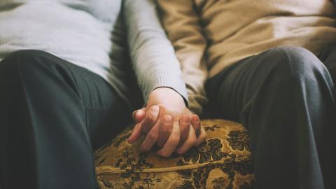 Zwei Menschen halten ihre Hände, während sie nebeneinander auf dem Sofa sitzen.