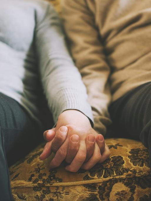 Zwei Menschen halten ihre Hände, während sie nebeneinander auf dem Sofa sitzen.