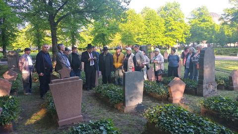 Eine Menschengruppe und ein Rabbiner stehen auf einem Friedhof vor Grabsteinen. 
