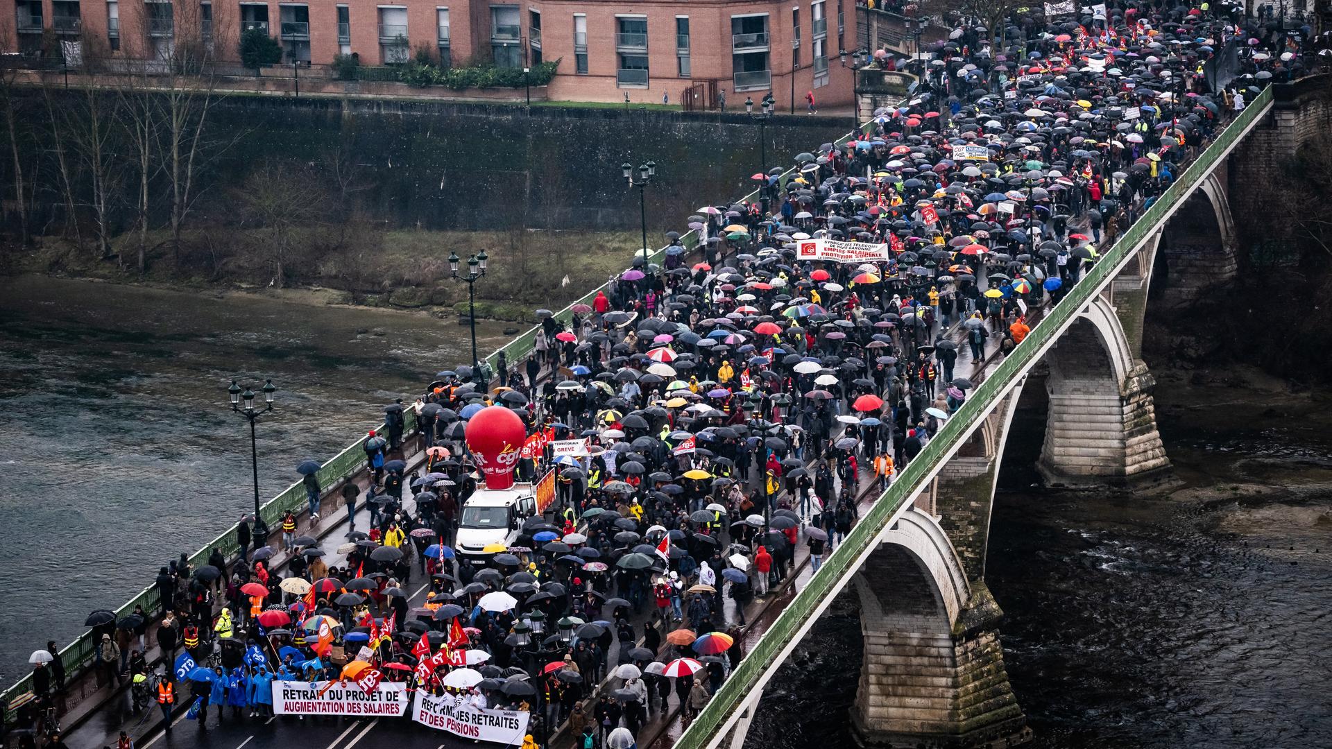 Das Foto zeigt eine Kundgebung gegen die Rentenreform in Toulouse. Viele Menschen mit Regenschirmen gehen über eine Brücke.