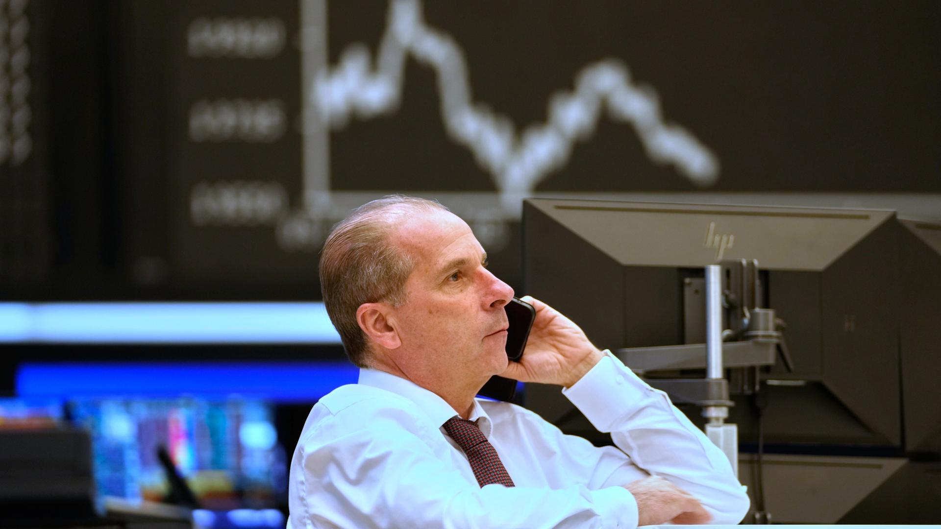 Ein Börsenmakler telefoniert. In Hintergrund wird auf einem Bildschirm der Stand des DAX angezeigt.