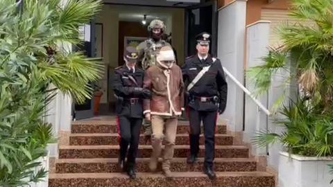Der Mafiaboss Matteo Messina Denaro wird von zwei Polizisten eine Treppe hinabgeführt, hinter ihnen ein schwer bewaffneter Soldat. 