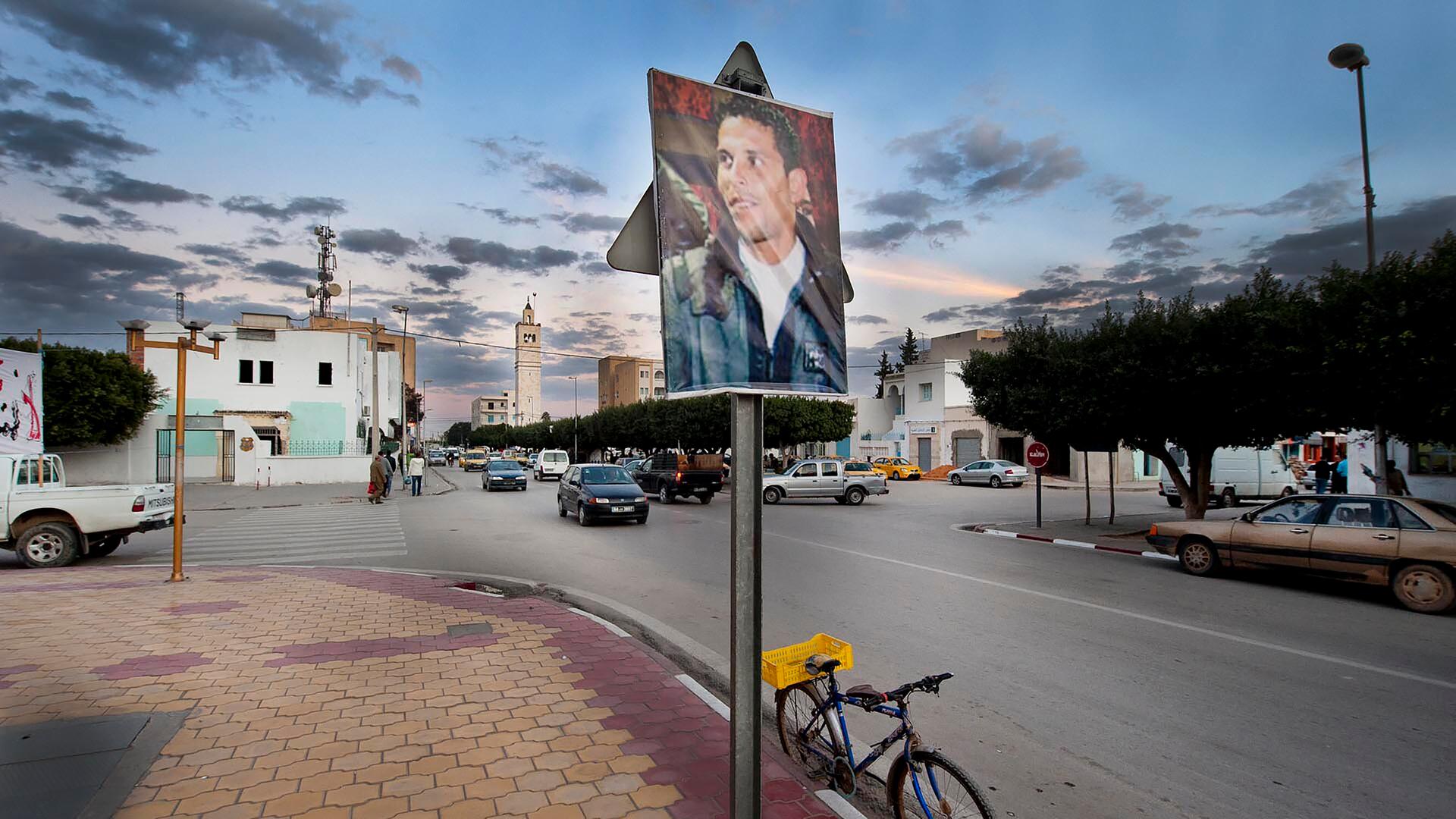 Ein Bild von Mohamed Bouazizi hängt außerhalb eines Regierungsgebäudes in Sidi Bouzid in Tunesien im Dezember 2011. Im Hintergrund sind Autos und alltägliches Leben zu sehen.