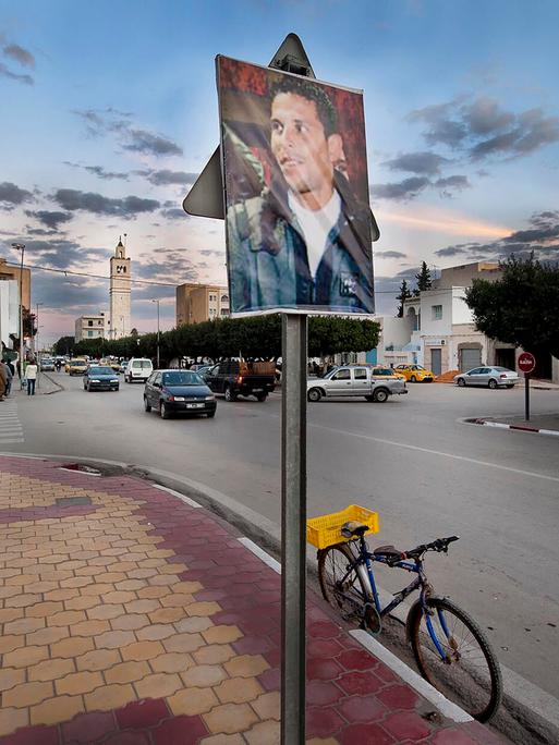 Ein Bild von Mohamed Bouazizi hängt außerhalb eines Regierungsgebäudes in Sidi Bouzid in Tunesien im Dezember 2011. Im Hintergrund sind Autos und alltägliches Leben zu sehen. Ein Bild von Mohamed Bouazizi hängt außerhalb eines Regierungsgebäudes in Sidi Bouzid in Tunesien im Dezember 2011. Im Hintergrund sind Autos und alltägliches Leben zu sehen.