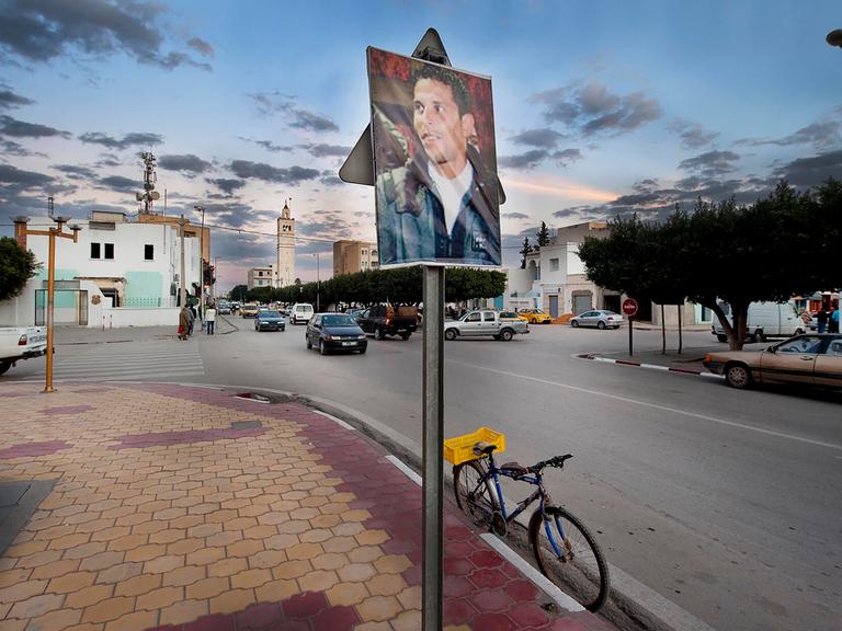 Ein Bild von Mohamed Bouazizi hängt außerhalb eines Regierungsgebäudes in Sidi Bouzid in Tunesien im Dezember 2011. Im Hintergrund sind Autos und alltägliches Leben zu sehen. Ein Bild von Mohamed Bouazizi hängt außerhalb eines Regierungsgebäudes in Sidi Bouzid in Tunesien im Dezember 2011. Im Hintergrund sind Autos und alltägliches Leben zu sehen.