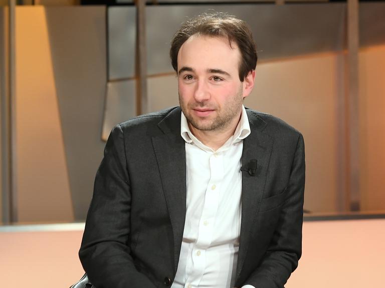 Ein Mann in weißem Hemd und dunklem Sakko sitzt als Gast in einer TV-Sendung. 