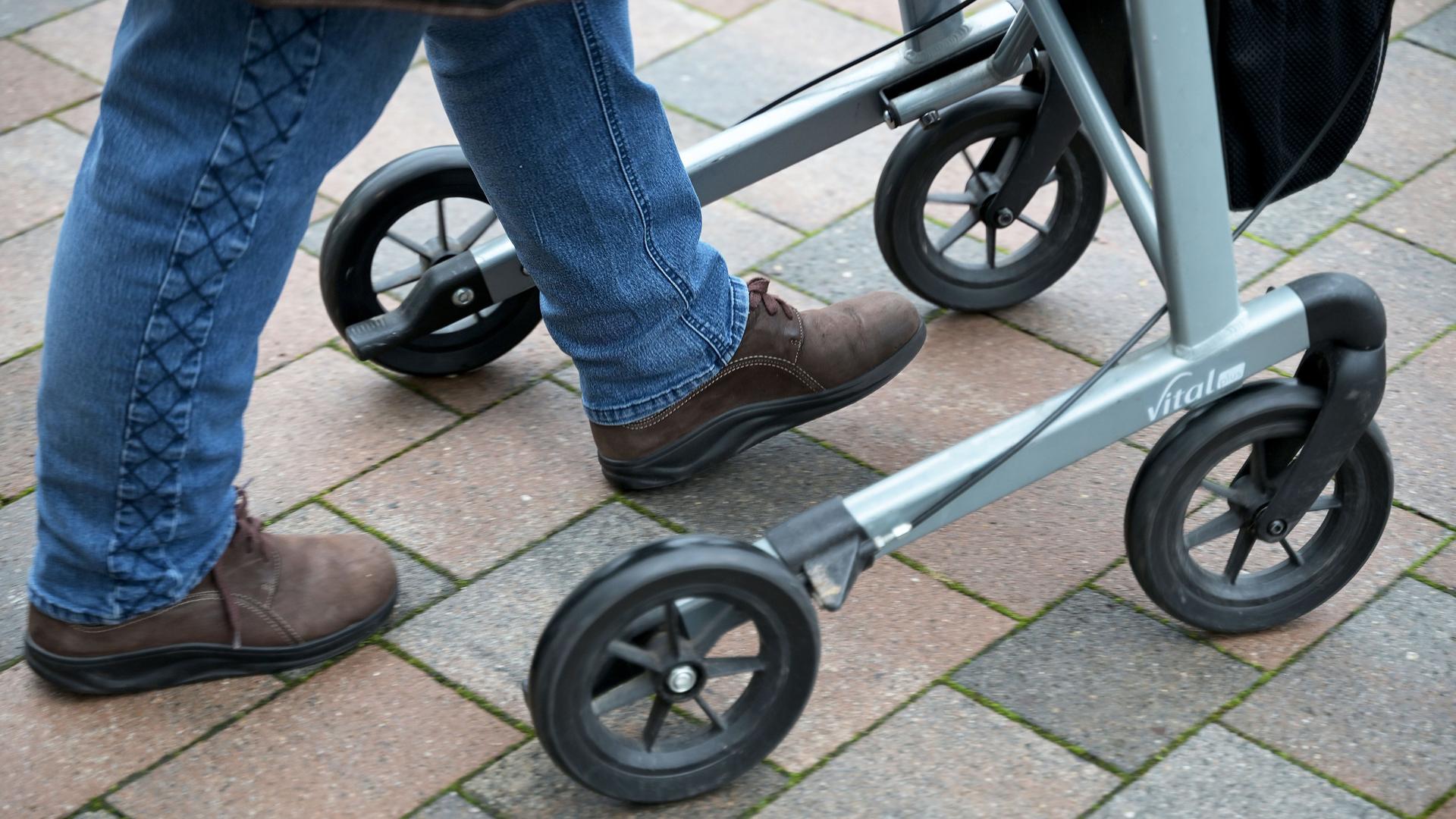 Eine pflegebedürftige Seniorin fährt mit ihrem Rollator auf einem gepflasterten Weg. Man sieht nur ihre Füße. (Symbolbild)