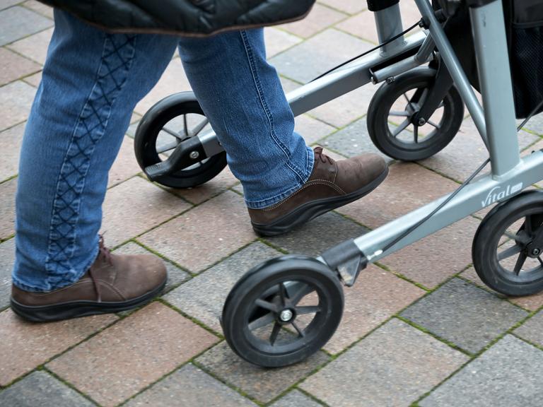 Eine pflegebedürftige Seniorin fährt mit ihrem Rollator auf einem gepflasterten Weg. Man sieht nur ihre Füße. (Symbolbild)