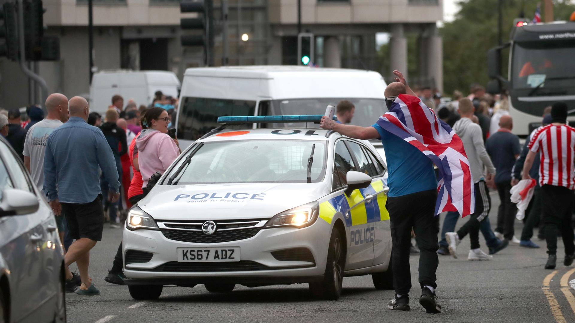 Ein Polizeiwagen steht inmitten von Demonstranten, ein Mann mit umgehängter britischer Flagge bewegt sich gestikulierend auf das Fahrzeug zu