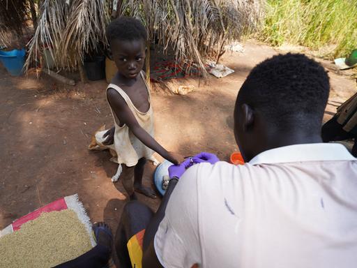 Ein kleiner Junge in einem Dorf trägt ein zerrissenes, dünnes Hemd und gibt seinen Daumen einem Mann, der eine Blutprobe entnimmt.