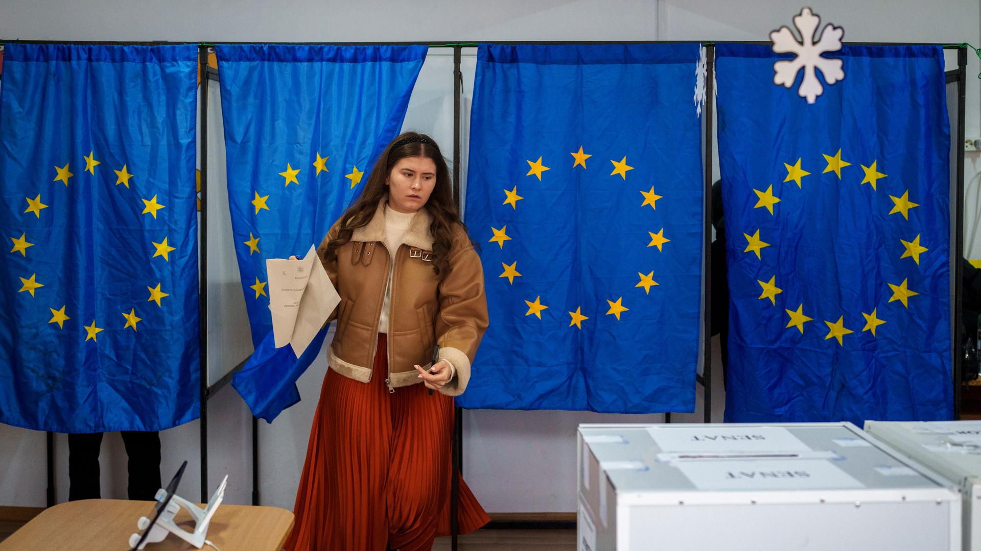 Eine Frau kommt aus der Wahlkabine, die mit EU-Flaggen abgetrennt sind. Sie geht zur Wahlurne. Rumänien wählt ein neues Parlament (1.12.2024) (AP Photo/Vadim Ghirda)