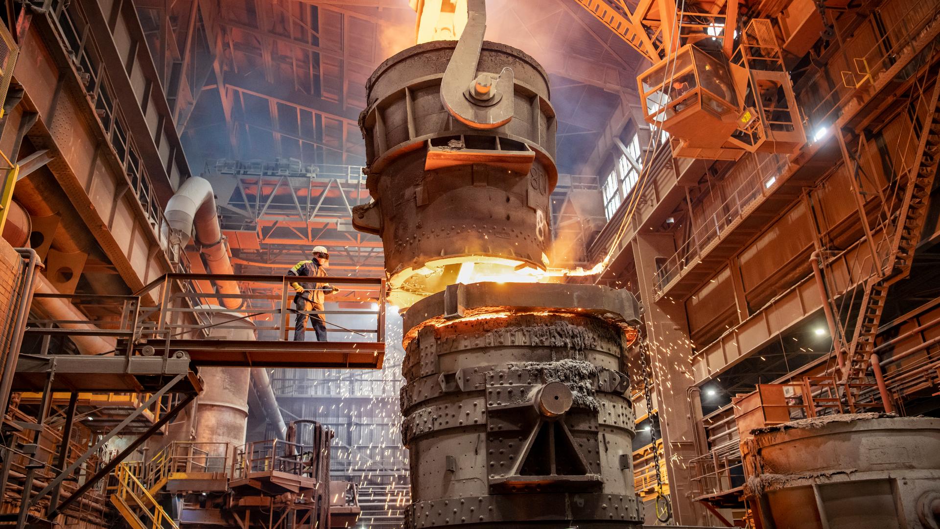 Ein Arbeiter im Schutzanzug beginnt mit dem Gießen von geschmolzenem Stahl im Stahlwerk in einer riesigen Produktionshalle.