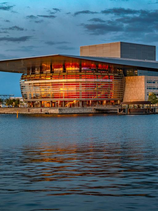 Die Königliche Oper in Kopenhagen in der Abenddämmerung, rötliches Licht scheint aus dem Foyer und spiegelt sich im Wasser.