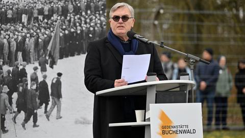 Hape Kerkeling steht an einem Rednerpult. Er trägt einen dunklen Mantel und eine Sonnenbrille. Hinter ihm steht ein schwarz-weißes Foto.