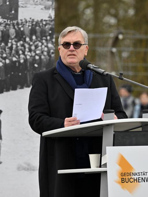 Hape Kerkeling steht an einem Rednerpult. Er trägt einen dunklen Mantel und eine Sonnenbrille. Hinter ihm steht ein schwarz-weißes Foto.