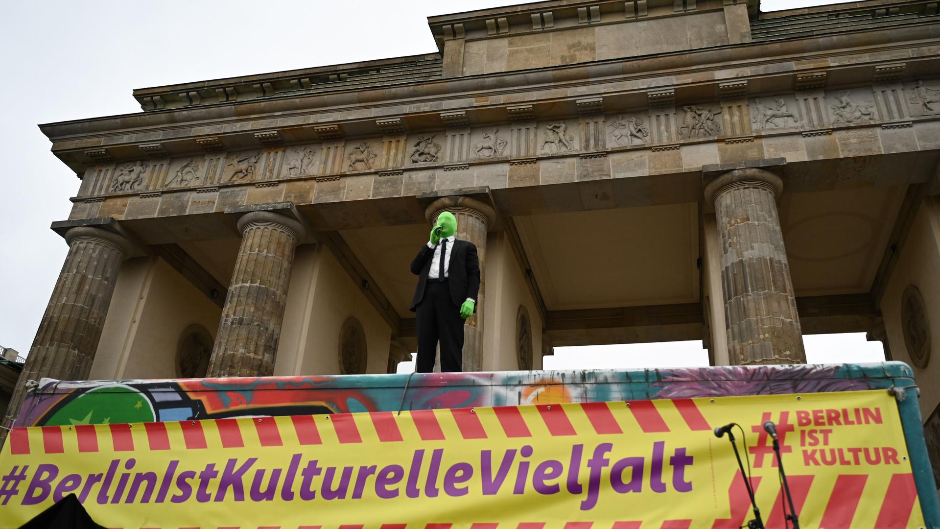 Das Foto zeigt den Schauspieler Lars Eidinger mit einer grünen Strumpf-Maske am Brandenburger-Tor.