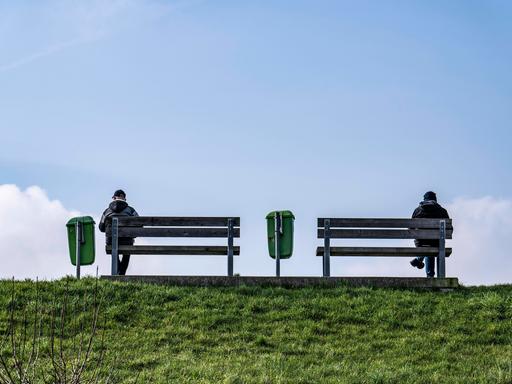 Zwei Personen sitzen bei blauem Himmel auf Sitzbänken auf einem Deich an der Weser in Bremerhaven.