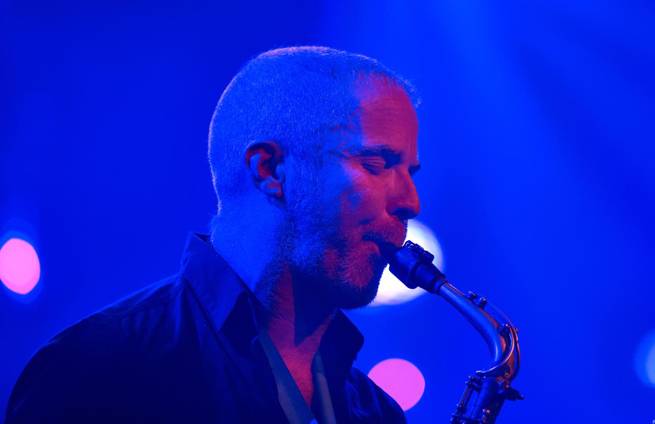 Ein Saxophonist mit extrem kurzen Haaren spielt sein Instrument, während er von blau-lila-farbenem Bühnenlicht umgeben ist.