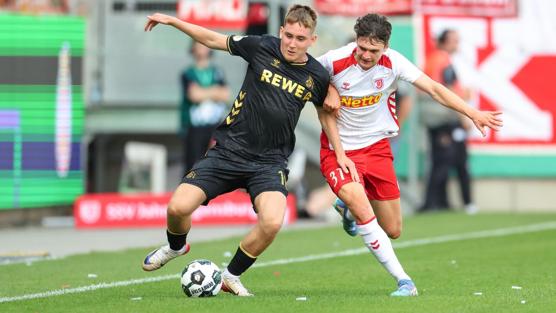 Das Bild zeigt einen Zweikampf von Fußballspielern auf dem Rasen eines Stadions in der Nähe einer Außenlinie.