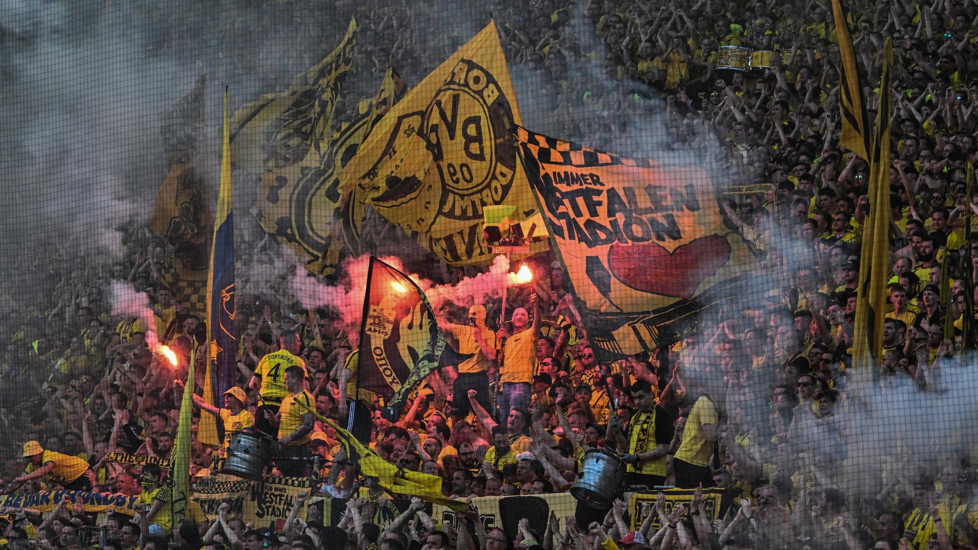 Gelb-schwarz gekleidete Fans auf den Rängen während einer Fußballspiels im Dortmunder Stadion. Manche schwenken fahnen, andere halten brennende Bengalos.