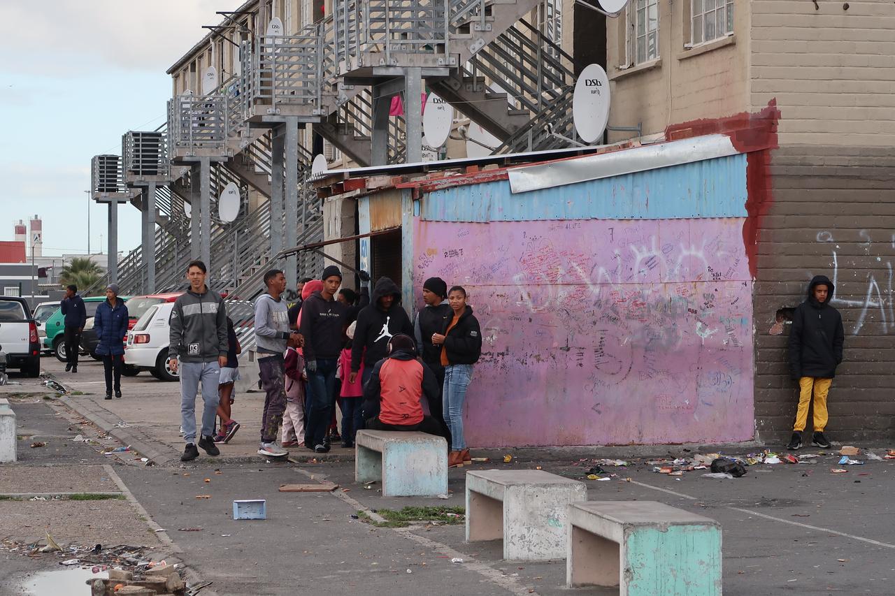 Blick auf eine Straße mit Sozialbauten. Außen an den Häusern sind Metalltreppen angebracht. Davor stehen schwarze Jugendliche in einer Gruppe herum.