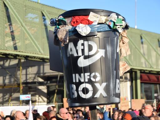 Auf einer Demonstration gegen Rechtsextremismus wird eine schwarze Tonne mit einem AfD-Logo hochgehalten. Die Tonne ist bis zum Rand gefüllt mit Müll.