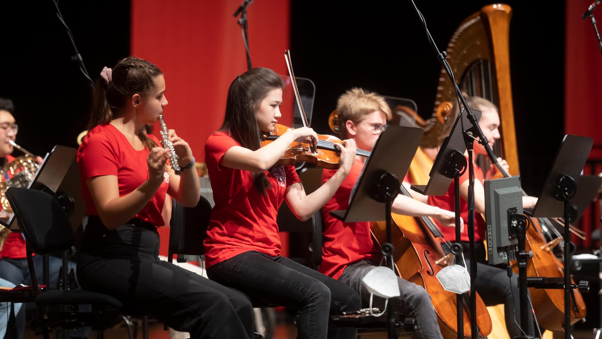 Junge Menschen sitzen auf Stühlen, halten ihre Instrumente und blicken in die Noten, die auf Notenständern vor ihnen stehen. Sie tragen alle rote T-Shirts und schwarze Hose. Zusammen bilden sie das Campus-Orchester beim Beethovenfest Bonn 2021.