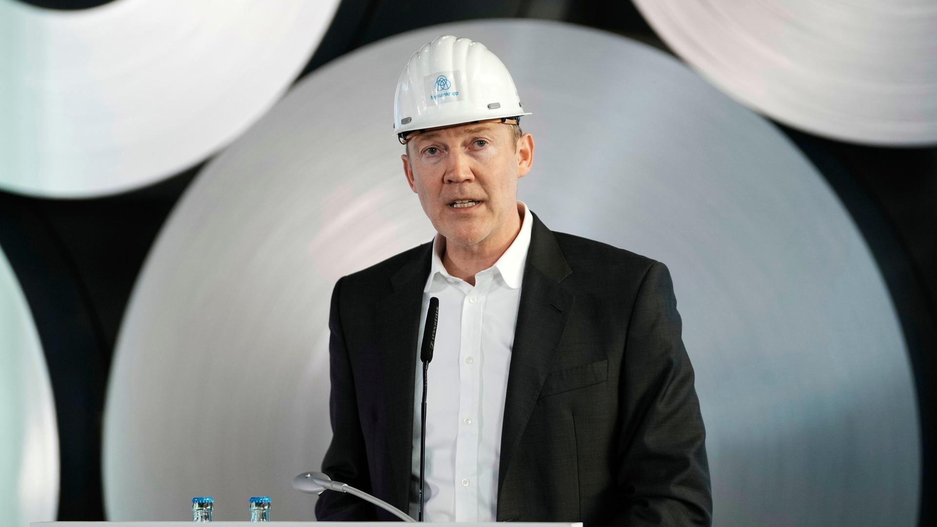 Dennis Grimm steht mit einem weißen Helm bei einer Veranstaltung hinter einem Rednerpult. Dennis Grimm steht mit einem weißen Helm bei einer Veranstaltung hinter einem Rednerpult.